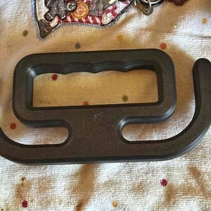 May include: A dark gray, rectangular handle with a central grip and curved extensions on either side. The handle appears to be made of a solid, possibly plastic or metal, material. The design suggests it is intended for carrying or lifting.