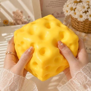 May include: A large, bright yellow, cube-shaped stress ball designed to resemble a block of Swiss cheese. The stress ball has numerous round holes and is held in two hands. The background includes a teddy bear and a basket of white flowers.