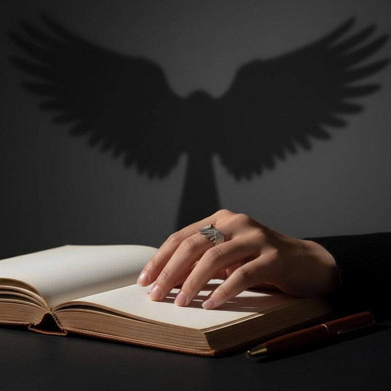 K&ouml;nnte beinhalten: Ein silberner Ring mit Fl&uuml;geldesign an einer Hand, die auf einem offenen Buch ruht. Ein brauner Stift liegt neben dem Buch. Der Schatten eines Vogels mit ausgebreiteten Fl&uuml;geln wird auf die Wand hinter der Hand und dem Buch geworfen.