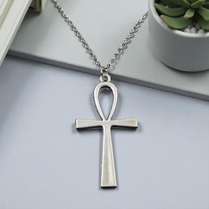 Könnte beinhalten: Halskette mit einem silberfarbenen Ankh-Anhänger. Das Ankh, ein Symbol des Lebens, hängt an einer Kette. Der Anhänger hat eine Schlaufe oben, einen horizontalen Balken und einen vertikalen Balken. Die Halskette wird auf einer hellgrauen Oberfläche präsentiert.