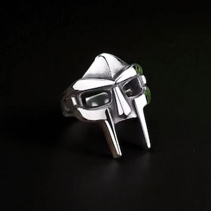 May include: A silver-colored ring in the shape of a mask, with a detailed design featuring eye and mouth openings. The ring has a polished finish and is set against a dark background.