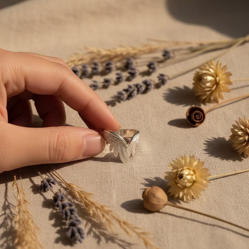 K&ouml;nnte beinhalten: Ein silberner Ring mit Blattdesign wird von einer Hand gehalten. Der Ring hat eine dreieckige Form mit detaillierten Bl&auml;ttermustern. Getrocknete Blumen und Lavendelzweige sind auf einer beigen Oberfl&auml;che um den Ring herum verstreut.