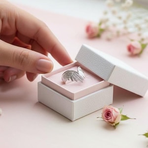 K&ouml;nnte beinhalten: Ein silberner Ring in Form von Engelsfl&uuml;geln liegt in einer ge&ouml;ffneten wei&szlig;en Schmuckschatulle mit rosa Innenraum. Eine Hand greift nach dem Ring. Rosa Rosen und kleine wei&szlig;e Bl&uuml;ten sind um die Schatulle verstreut.