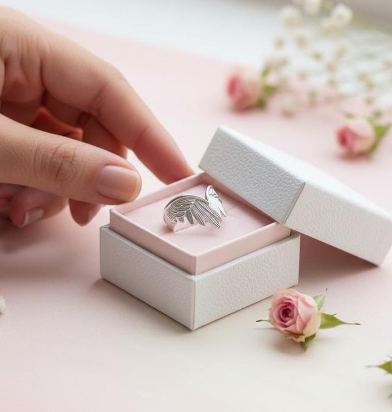 K&ouml;nnte beinhalten: Ein silberner Ring in Form von Engelsfl&uuml;geln liegt in einer ge&ouml;ffneten wei&szlig;en Schmuckschatulle mit rosa Innenraum. Eine Hand greift nach dem Ring. Rosa Rosen und kleine wei&szlig;e Bl&uuml;ten sind um die Schatulle verstreut.