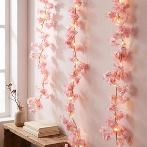Peut inclure: Trois guirlandes verticales de fleurs de cerisier artificielles dans des tons rose clair, chacune entrelacée de fines lumières LED blanc chaud, suspendues contre un mur uni pour créer une décoration florale lumineuse.