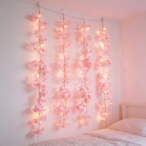 May include: Pink cherry blossom string lights hanging on a white wall. The lights are strung along a thin wire, with the blossoms cascading down. The room is painted in a soft pink hue, creating a dreamy atmosphere.