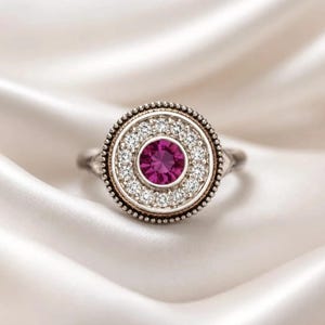 Puede incluir: Un anillo de tono plateado con una cara circular y una piedra preciosa magenta de corte redondo en el centro. La piedra central está rodeada por un halo de pequeñas piedras blancas transparentes de corte redondo y un borde metálico perlado.