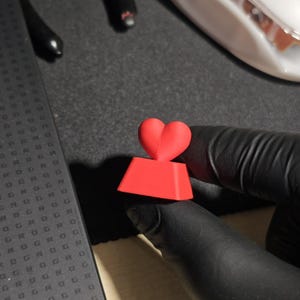 May include: A red heart-shaped keycap with a square base. The keycap is made of plastic and has a smooth surface.