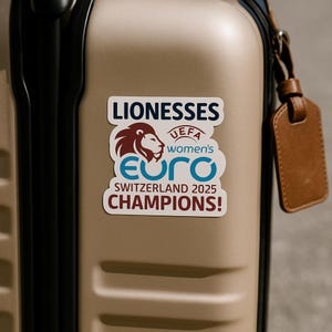 May include: A sticker on a tan suitcase displays the text "LIONESSES" above a logo with a lion's head and the words "UEFA women's EURO Switzerland 2025 CHAMPIONS!" in white and blue. A brown leather luggage tag hangs from the handle.