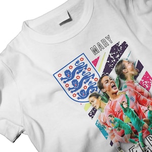 Puede incluir: Camiseta blanca con un diseño gráfico que presenta el escudo del equipo nacional de fútbol de Inglaterra e imágenes de jugadoras de fútbol. La palabra "MARY" está impresa encima de las imágenes. El diseño incorpora acentos azules, rosas, verdes y morados.