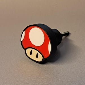 May include: A mushroom-shaped cabinet knob with a red cap, white spots, and a black base. The knob features a cartoon-style design with a smiling face. The knob is a decorative element for furniture.