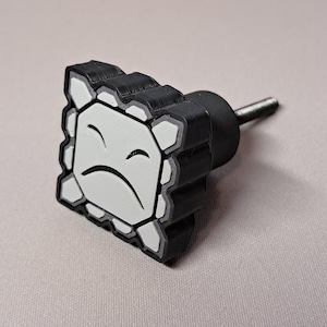 May include: A black and white cabinet knob with a cartoon face design. The knob features a sad face with a white background and a black outline. The knob is made of a durable material and has a metal screw for easy installation.