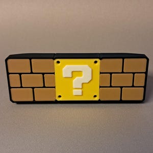May include: A rectangular novelty item resembling a brick wall from a video game. The item features a yellow square with a white question mark in the center, surrounded by brown brick-like shapes and a black border.