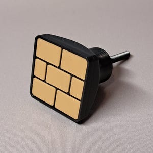May include: A black cabinet knob with a square face featuring a brick pattern in tan and black. The knob has a cylindrical post for mounting. The design is modern and suitable for various furniture styles.