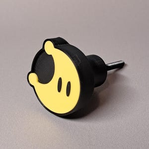 May include: A black and yellow cabinet knob with a cartoon-like design. The knob features a circular face with a yellow crescent shape and two black oval eyes. The knob's body is black, and it has a silver-colored screw.