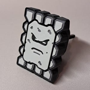 May include: A black and white Thwomp character from the Super Mario video game series. The Thwomp is a rectangular block with a grumpy face. The edges are black, and the face is white with black details. The Thwomp has a black post on the back.