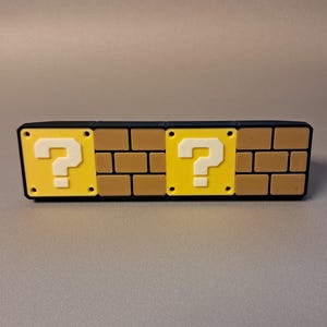 May include: A rectangular, brick-patterned object with two yellow blocks featuring white question marks. The object has a black border and is likely a decorative item or a novelty product inspired by a video game.