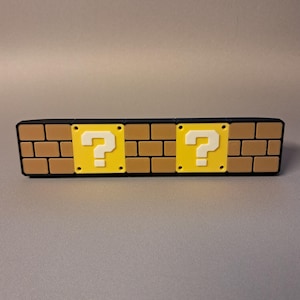 May include: A black rectangular object with a brick pattern and two yellow question mark blocks. The brick pattern is tan and brown. The question mark blocks are yellow with a white question mark.