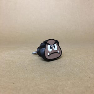 May include: A brown and black 3D printed door knob shaped like a Goomba from the Super Mario Bros. video game series. The Goomba has a frowning face with white eyes and teeth.
