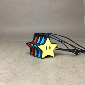 May include: A collection of five star-shaped pendants in red, green, blue, and yellow. Each pendant has a black border and is strung on a black cord. The yellow star has two black dots for eyes.