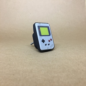 May include: A black and white handheld video game console shaped like a Game Boy with a green screen and red buttons. The console is mounted on a white knob.