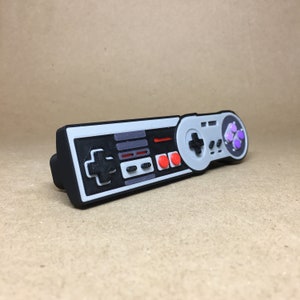 May include: A gray and black plastic video game controller with red and purple buttons. The controller is shaped like a classic NES controller with a D-pad and four face buttons.