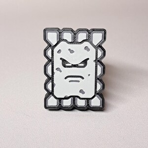 May include: A rectangular, black, white, and gray pin featuring a cartoon character with an angry expression. The character has a white face with gray spots, a black outline, and a gray border. The pin is a collectible item.