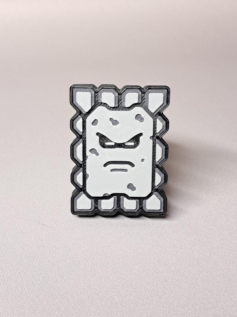 May include: A rectangular, black, white, and gray pin featuring a cartoon character with an angry expression. The character has a white face with gray spots, a black outline, and a gray border. The pin is a collectible item.