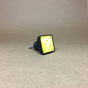 May include: A black and yellow question mark block knob. The knob is a 3D printed replica of a classic video game item.