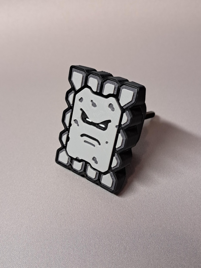 May include: A black and white Thwomp character from the Super Mario video game series. The Thwomp is a rectangular block with a grumpy face. The edges are black, and the face is white with black details. The Thwomp has a black post on the back.