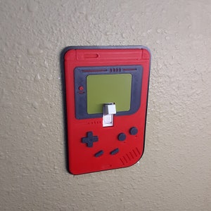 May include: A red and black light switch cover shaped like a vintage handheld video game console. The switch is labeled "ON".