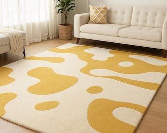 May include: A cream-colored area rug with a mustard yellow abstract pattern. The rug is rectangular and covers a wooden floor. A white sofa, a plant, and a gold lamp are in the background.