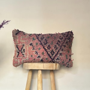 May include: A rectangular, textured pillow with a pink and purple color scheme. The pillow features a geometric pattern with dark blue and green accents. It is displayed on a small, wooden stool.