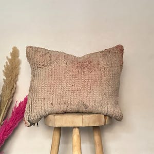 Puede incluir: Un cojín rectangular y texturizado en tonos beige y rosa descansa sobre un pequeño taburete de madera. El cojín tiene un aspecto tejido y un aspecto ligeramente desgastado. Plantas secas decorativas en negro, morado, beige y rosa a la izquierda.