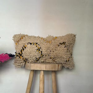 May include: A beige rectangular pillow with a textured, shaggy surface, adorned with black and yellow circular patterns. The pillow rests on a small, three-legged wooden stool. Decorative dried flowers in pink, beige, and black are in the background.