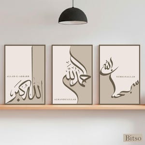 Puede incluir: Tres impresiones artísticas enmarcadas con caligrafía árabe en tonos marrón y beige. Cada impresión presenta una frase diferente, incluyendo "Allah-u-Akbar", "Alhamdulillah" y "Subhanallah". Las impresiones se exhiben en una estantería de color claro.