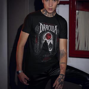 May include: A black short-sleeve t-shirt featuring a graphic print of Bram Stoker's Dracula. The design displays a silhouette of a figure in a cape standing before a gothic archway with a large red moon, bats, and text that reads I am Dracula.
