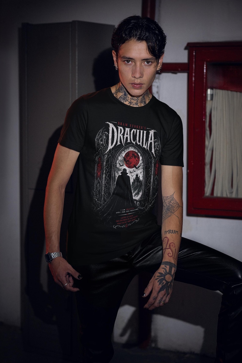 May include: A black short-sleeve t-shirt featuring a graphic print of Bram Stoker's Dracula. The design displays a silhouette of a figure in a cape standing before a gothic archway with a large red moon, bats, and text that reads I am Dracula.