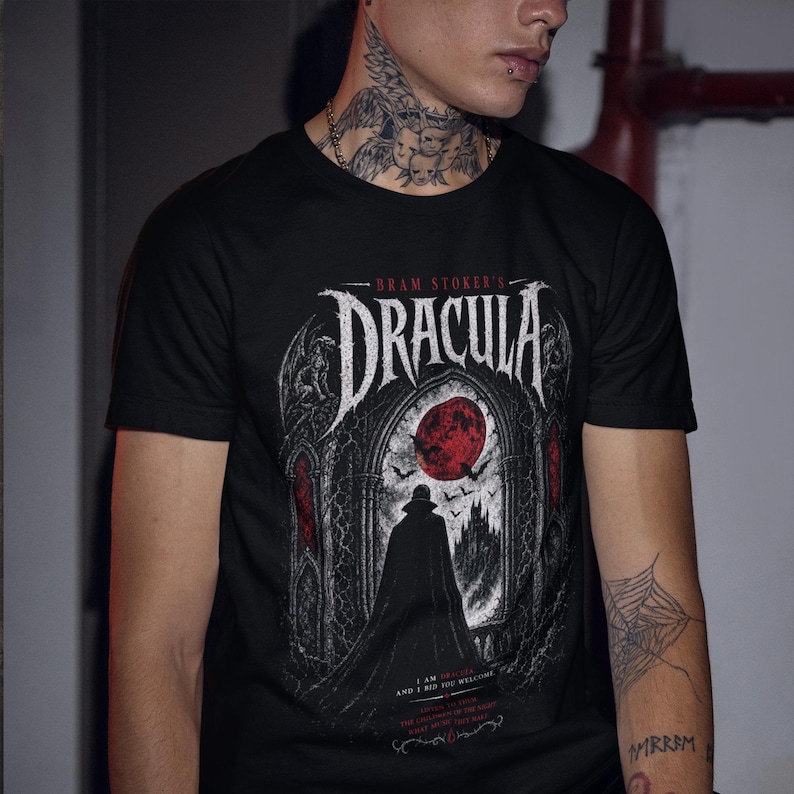 May include: A black short-sleeved t-shirt featuring a graphic print of Bram Stoker's Dracula. The design displays a cloaked figure standing before a gothic archway, a large red moon, bats, and a castle. The text reads BRAM STOKER'S DRACULA.