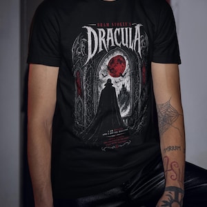 May include: A black short-sleeve t-shirt featuring a graphic print of Bram Stoker's Dracula. The design displays a silhouette of a figure in a cape standing before a gothic archway with a large red moon, bats, and text in white and red ink.