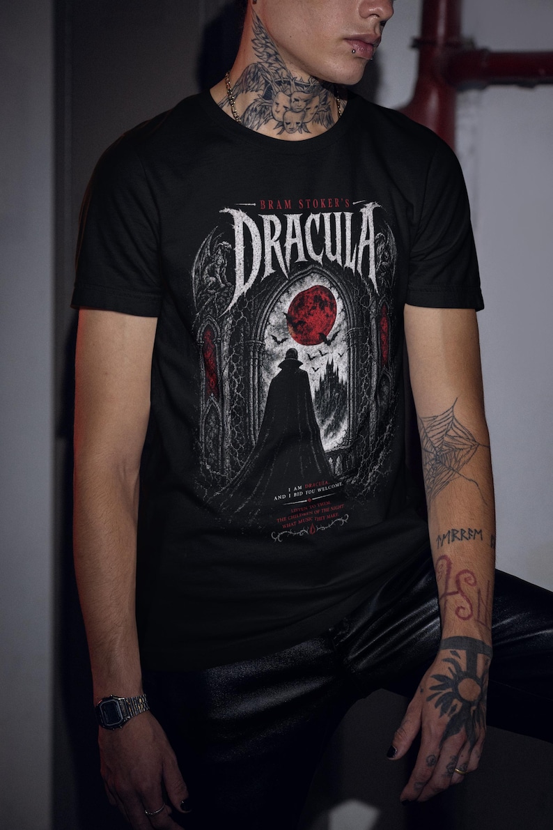 May include: A black short-sleeve t-shirt featuring a graphic print of Bram Stoker's Dracula. The design displays a silhouette of a figure in a cape standing before a gothic archway with a large red moon, bats, and text in white and red ink.