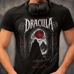 May include: A black short-sleeved t-shirt featuring a graphic print of Bram Stoker's Dracula. The design shows a figure in a cape standing before a gothic archway, a red moon, and a castle, with the text "I am Dracula, and I bid you welcome."