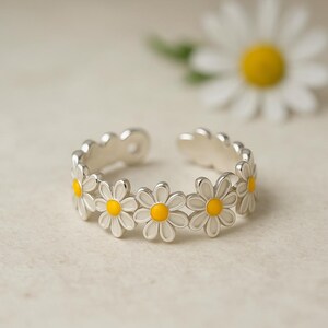 Puede incluir: Un anillo de margarita plateado con pétalos blancos y centros amarillos. El anillo ajustable presenta un diseño floral con múltiples flores de margarita. El anillo está abierto en la parte posterior y se encuentra sobre una superficie neutra.