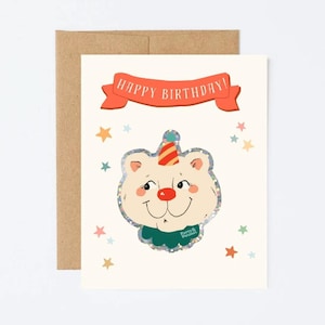 May include: A birthday card featuring a cartoon bear with a party hat and a red nose. The card has a banner that reads "HAPPY BIRTHDAY!" and is decorated with colorful stars. The card is paired with a brown envelope.