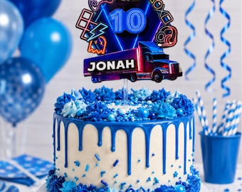 May include: A round cake with white frosting and blue icing drips, topped with a cake topper featuring a blue truck, the name JONAH, the number 10, and gaming icons with the text GAME OVER. The cake is decorated with blue frosting swirls.