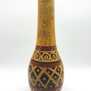 May include: A tall, slender vase with a rounded base and a narrow neck. The surface features a distressed yellow and brown finish with hand-painted black and dark red geometric patterns, including horizontal bands, circles, and a diamond grid.