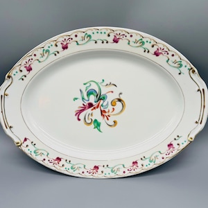 May include: An oval white porcelain serving platter featuring a central hand-painted floral motif in shades of magenta, teal, and gold. The rim is decorated with a matching scrolling vine pattern and gold-painted accents on the handles.