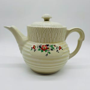 May include: A cream-colored ceramic teapot featuring a ribbed body and a lid with a matching finial. The upper section has a vertical leaf-like embossed pattern, while the center displays a colorful floral decal with red, yellow, and green.