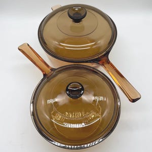 May include: Two amber-colored glass saucepans with matching glass lids. Each saucepan features a long, integrated glass handle and a circular lid with a central knob. The glass is translucent and tinted in a warm brown hue, showing a smooth finish.