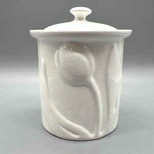 May include: A white ceramic canister with a lid and a tulip design. The canister is cylindrical with a slightly flared rim and a small knob on the lid. The tulip design is embossed on the canister's surface.